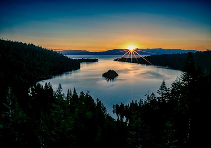 the sun sets over distant mountains as the viewer stands and looks at a forest rimmed bay in a large lake