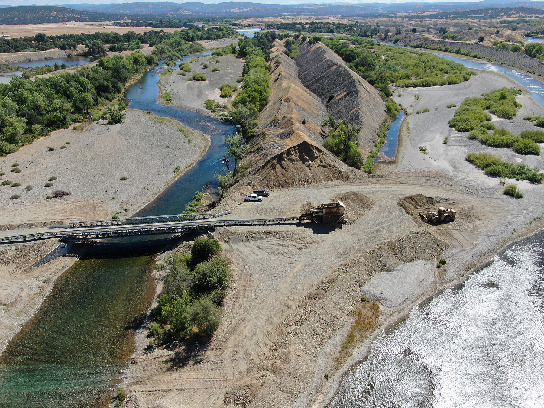 photograph showing land punctuated by meandering waterways, a bridge, construction equipment, earthen embankments, and trees