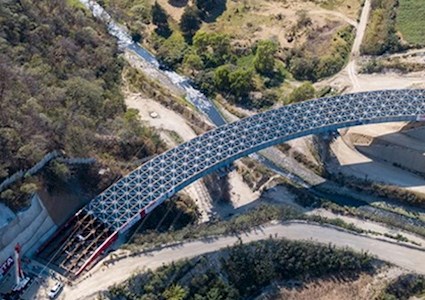 Aerial view of a large arched lattice bridge spanning a river with construction equipment near one bridge base.