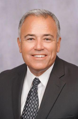 Portrait of Victor Fernandez-Cuervo, UESI President for 2025. Victor is a fair-skinned Hispanic man with gray hair. In this image, he is wearing a suit and tie in front of a gray background.