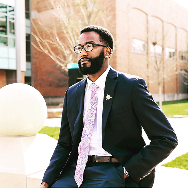 man in black suit with white shirt and multicolored tie. He is wearing black glasses and has an earring in his left ear.