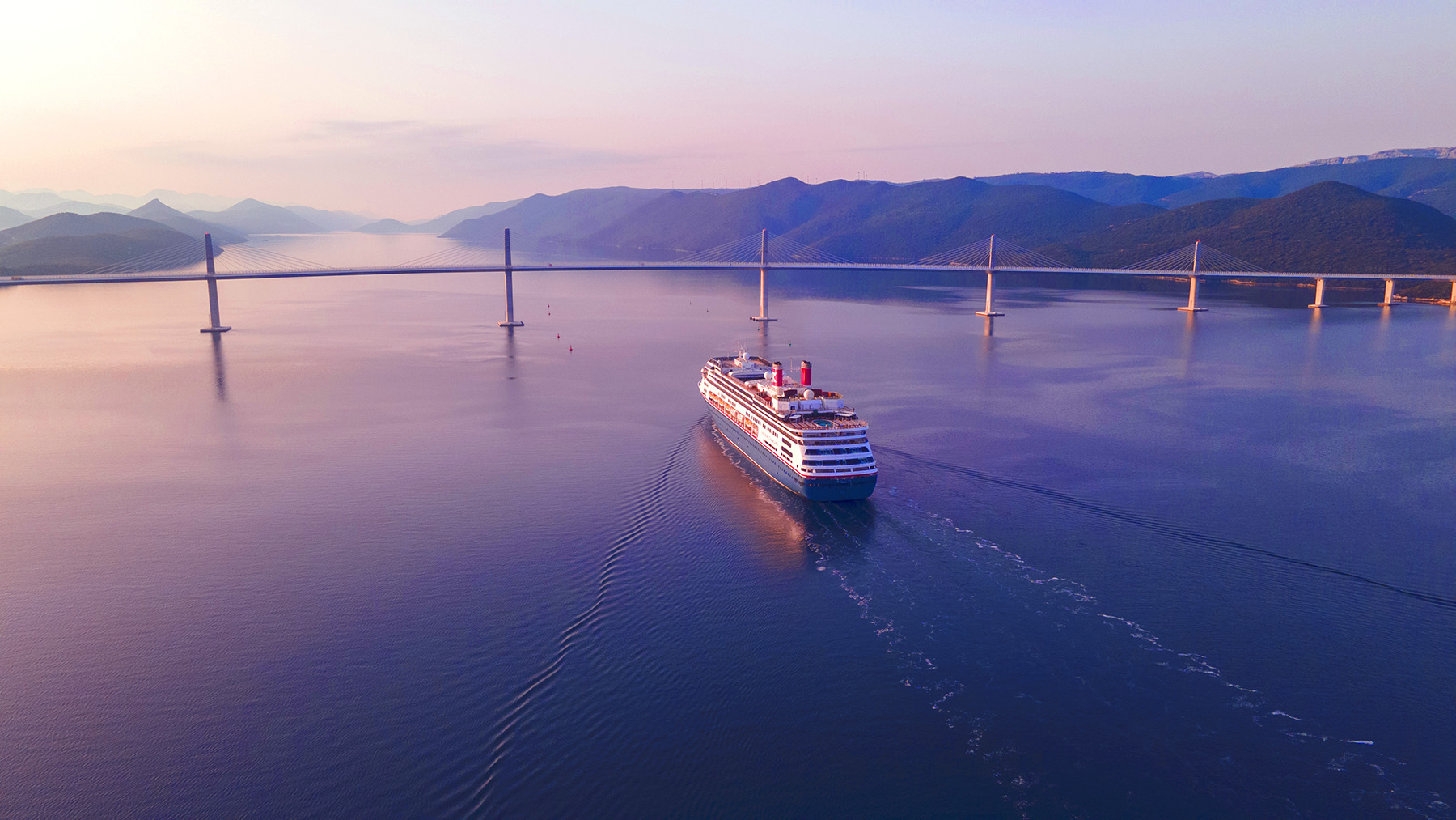 A wide-angle shot of the bridge with columns and cables that meet to form triangles. The water reflects pink and blue colors. In the background, there are a few small mountains. A barge floats in the foreground. 