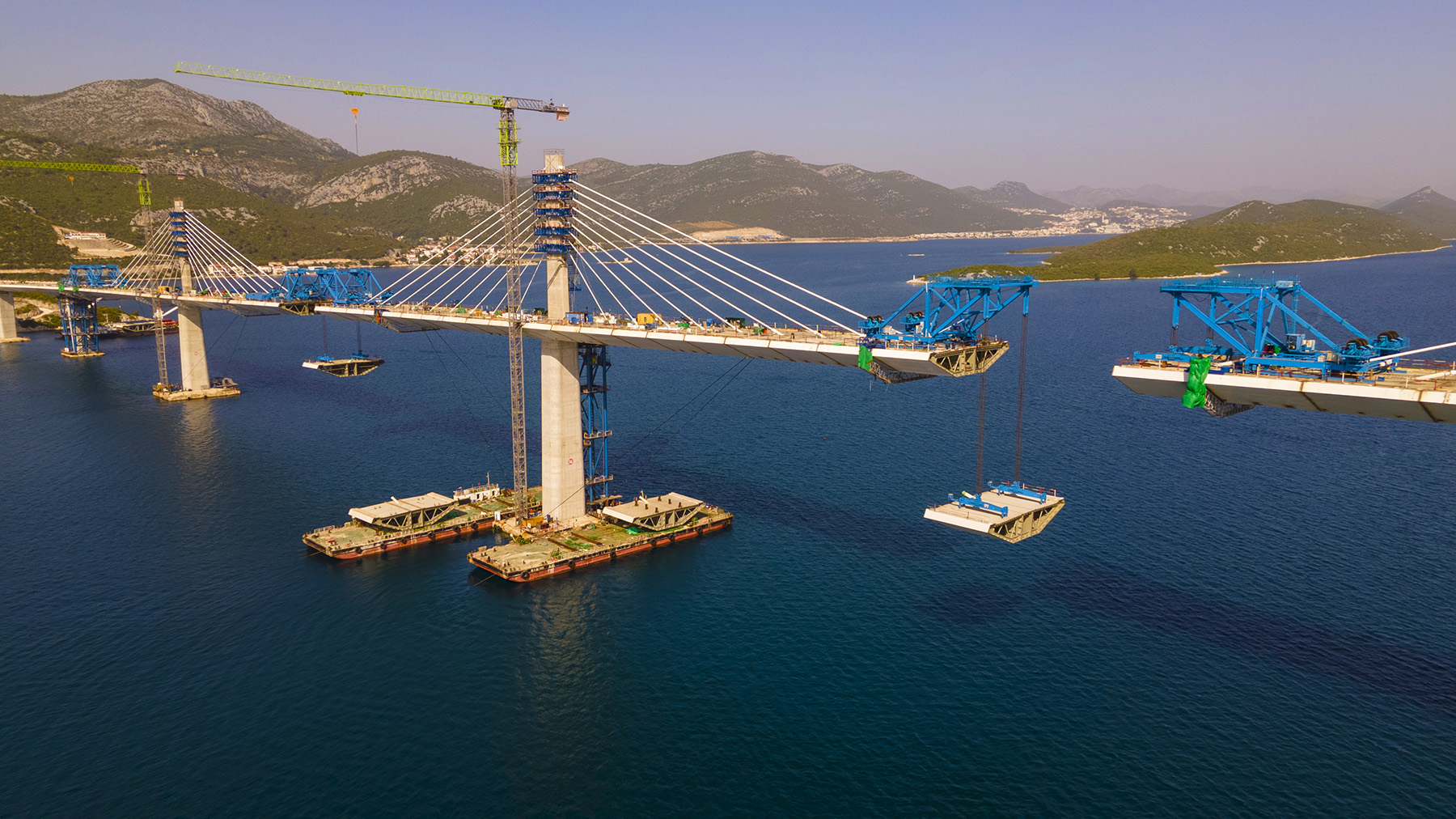 Barges are floating in the water next to one of the bridge’s supporting columns. Blue cranes sitting on the bridge deck lift middle sections onto the bridge. 