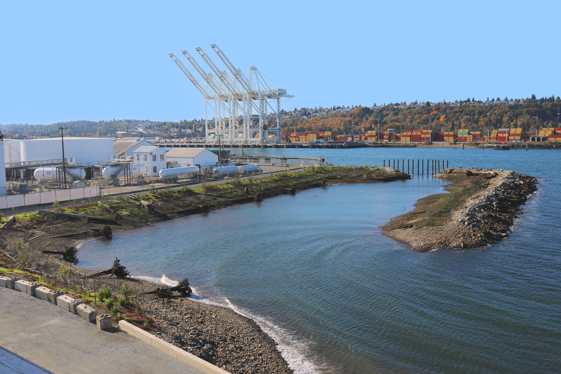 Photograph shows a body of water with industrial buildings on one side and a road on the other. In the water on the right is a structure made of earth and rocks with vegetation on it. 