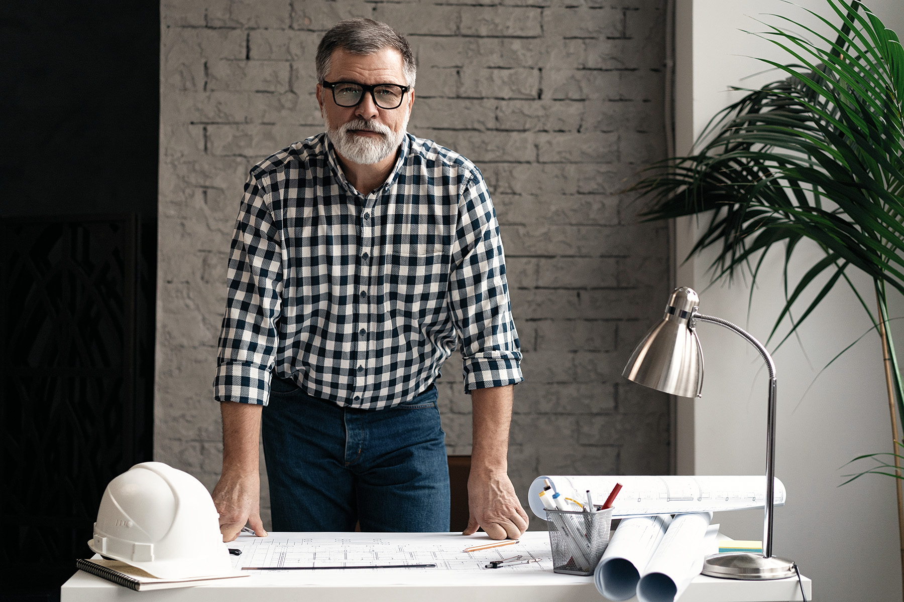 A middle-aged White man with a gray beard and black glasses, wearing a black-and-white-checkered button-down shirt leans over an architectural drawing on a table. 