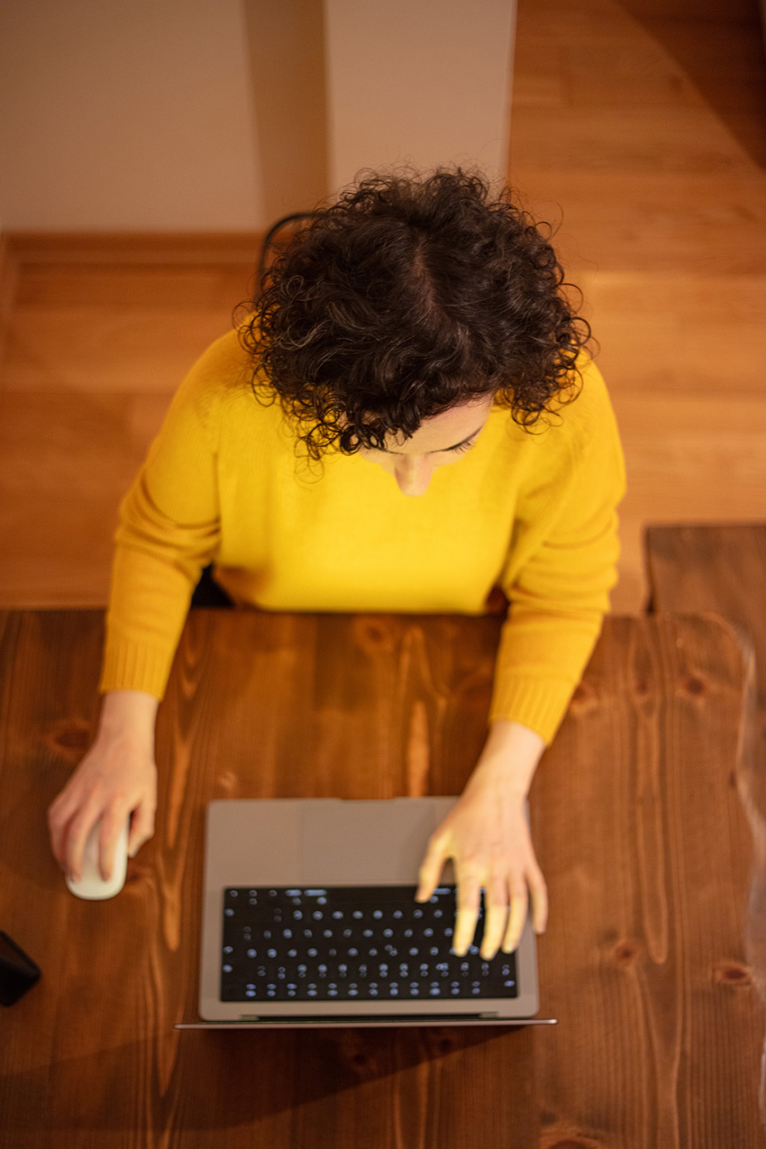 A woman with brown curly hair wearing a bright yellow sweater works on a laptop.
