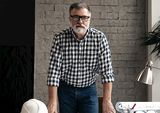A middle-aged White man with a gray beard and black glasses, wearing a black-and-white-checkered button-down shirt leans over an architectural drawing on a table.
