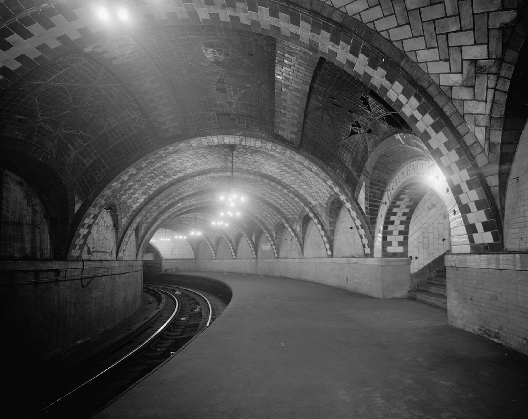 Photograph shows the interior of a subway station—its tracks and decorative ceiling tiles. 