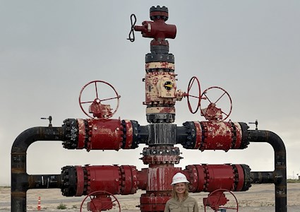 a woman in a construction hat stands in front of a red and black wellhead