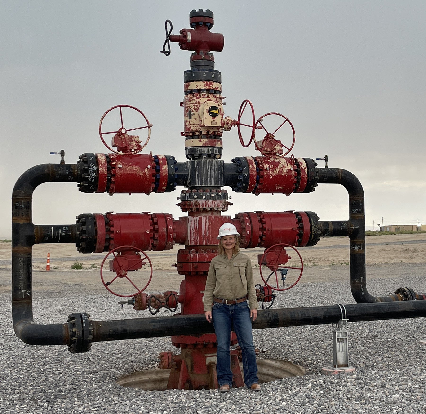 a woman wearing a hardhat stands in front of a red and black wellhead