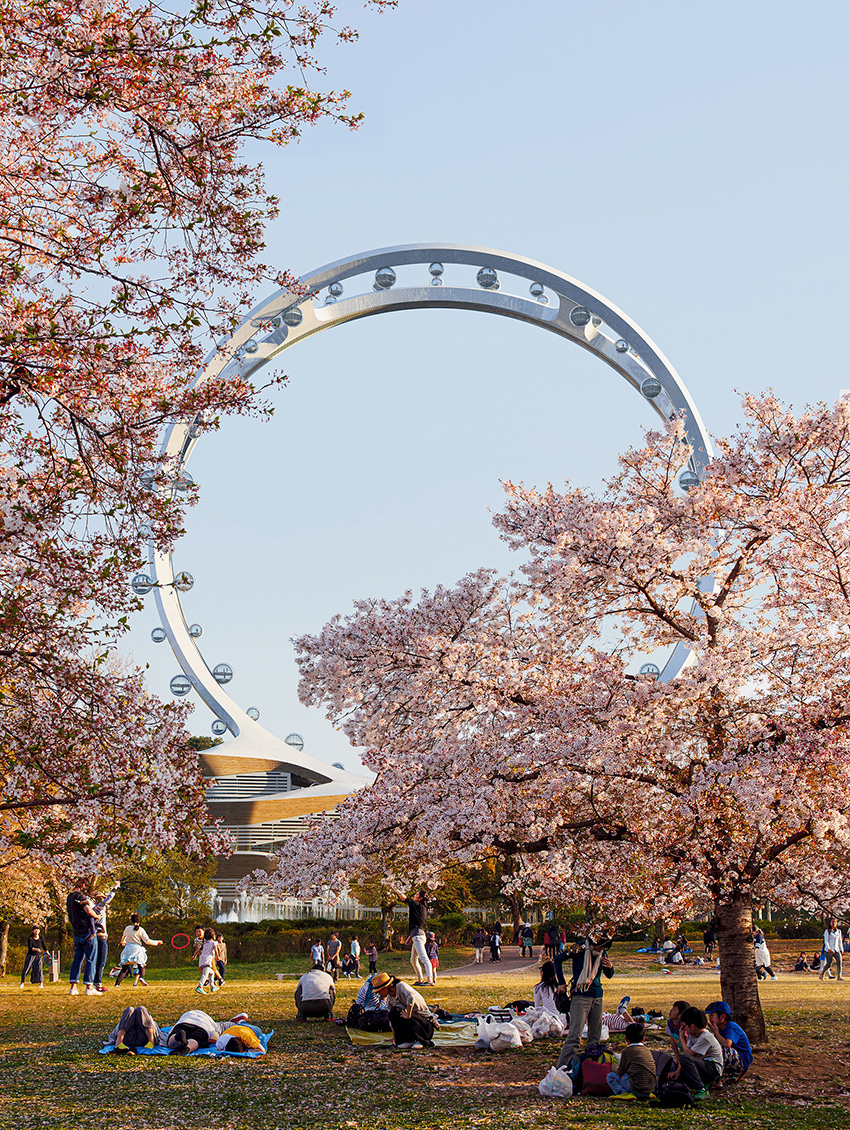 Rendering shows an intersecting-ring, dual-track, spokeless Ferris wheel in the background. In the foreground, people walk through a park and lounge on the grass in the park. 