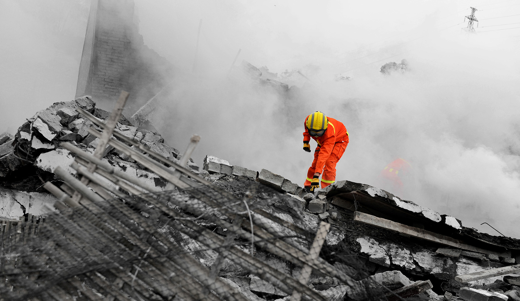 A first responder in full orange personal protective gear and thick goggles stands on a mound of collapsed structure. 