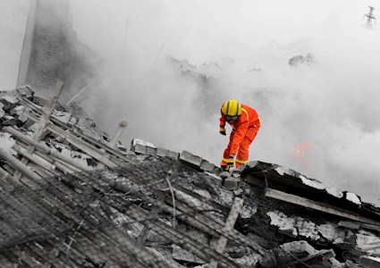 A first responder in full orange personal protective gear and thick goggles stands on a mound of collapsed structure. 