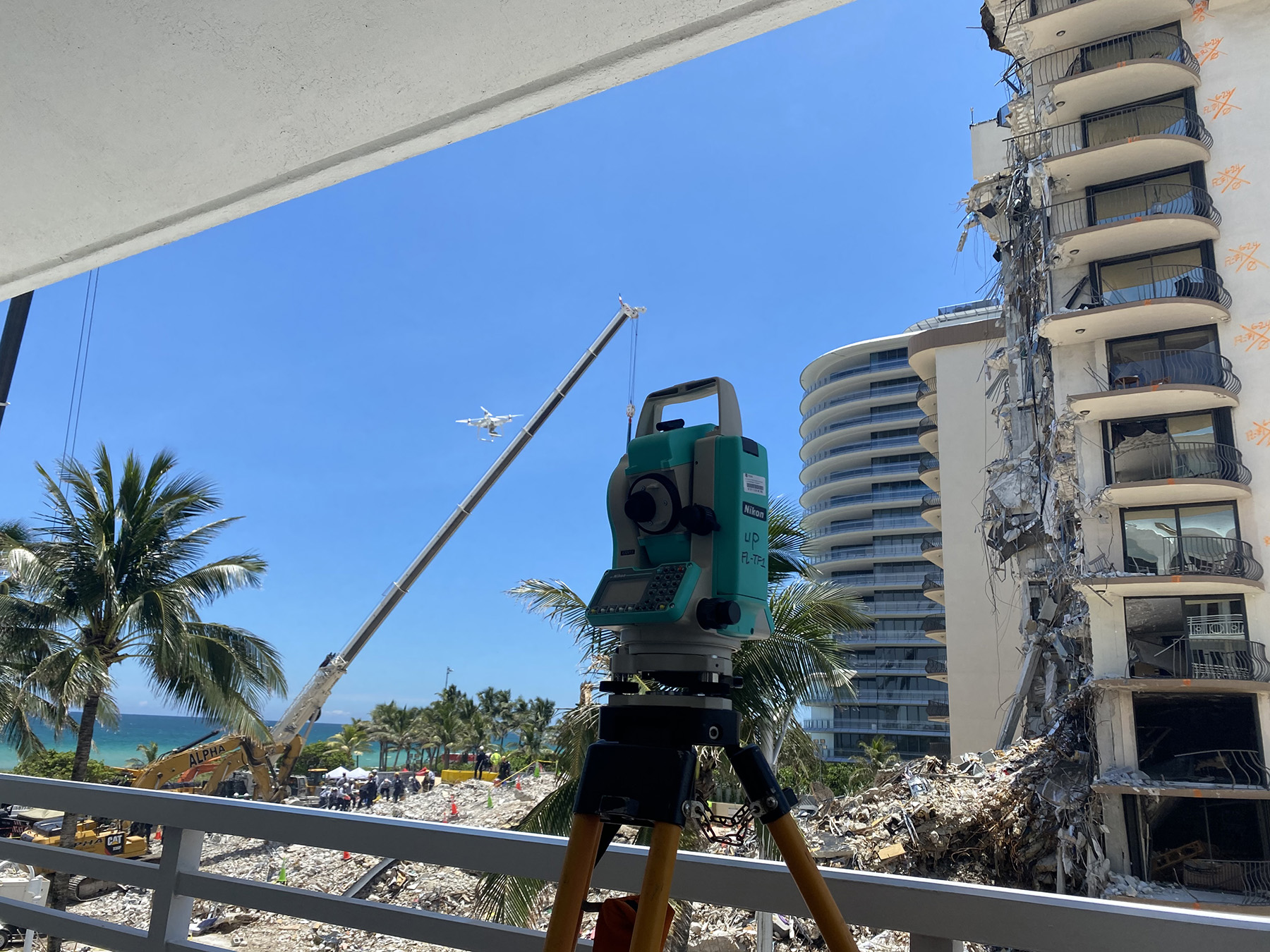 In the foreground, a turquoise survey telescope standing on four legs. In the background, a partially collapsed building to the left, and people standing on rubble to the right. Adjacent the rubble are palm trees and the Atlantic Ocean. 