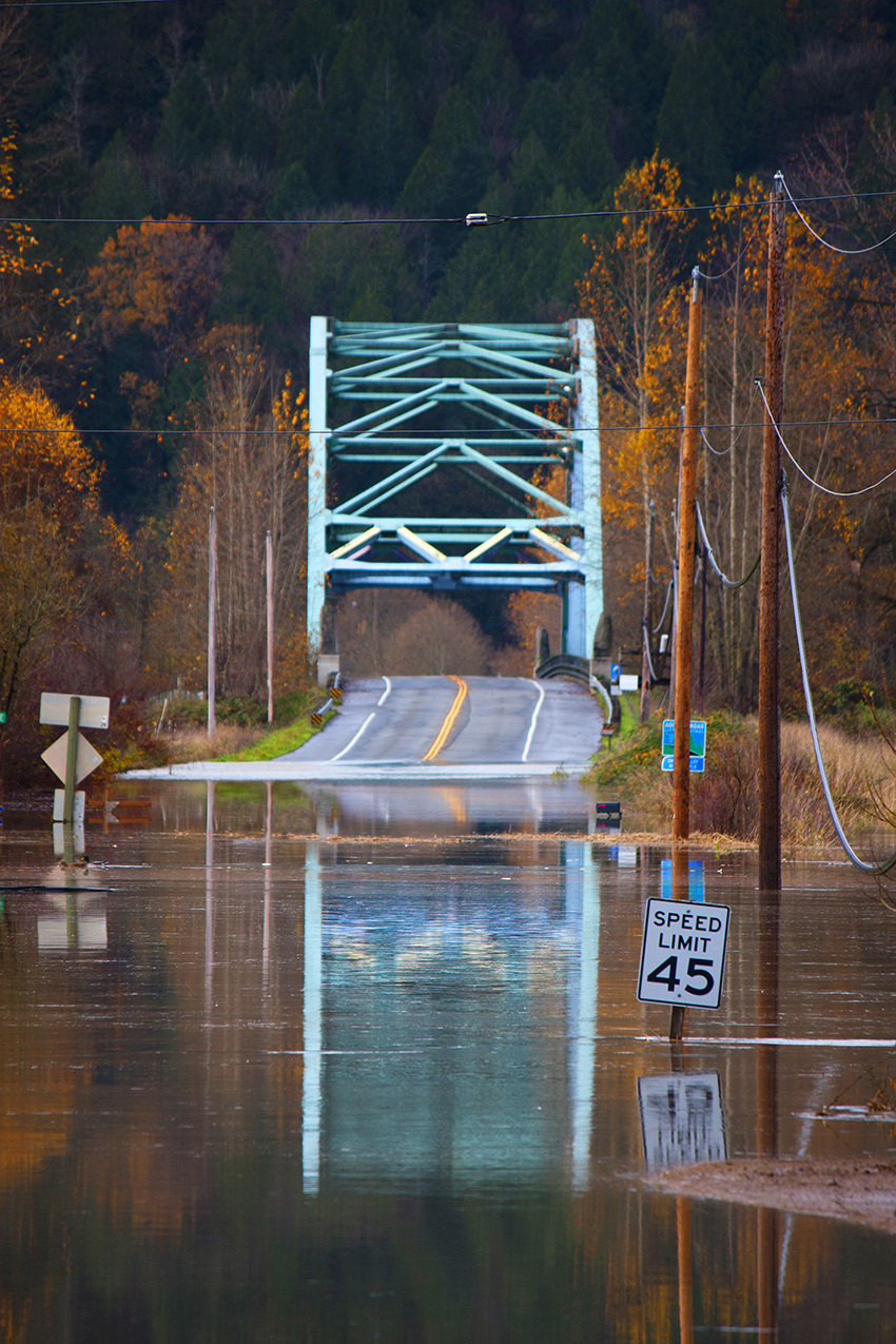 The photograph shows a flooded road on the approach to a steel bridge. 