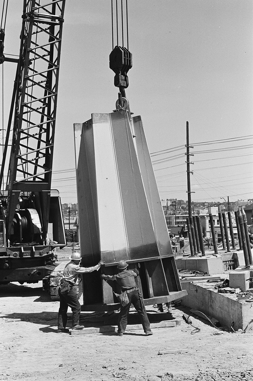 Two men move a large steel cylindrical object into a concrete and steel base. 