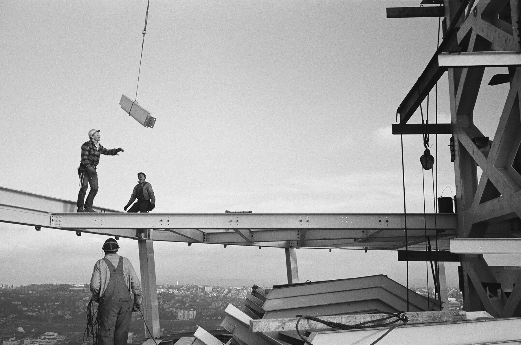 Three men stand on steel beams. One has his hands raised, waiting to catch a steel piece as a crane swings it toward him. 