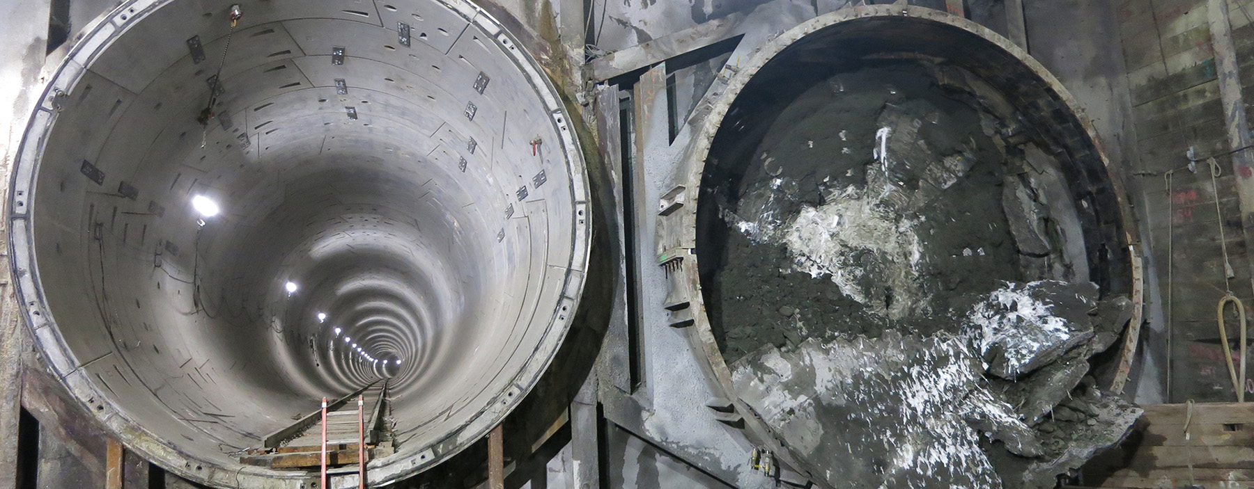 Two massive tunnels are side by side underground. On the left is an open tunnel with a track along the bottom; on the right, is a tunnel filled with soil. 