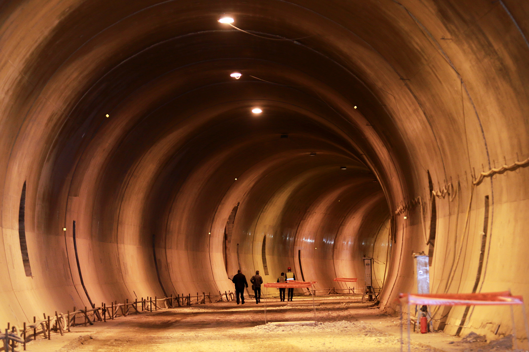 Four workers, two with reflective yellow vests, are pictured walking from behind in an underground tunnel with orange lighting. 