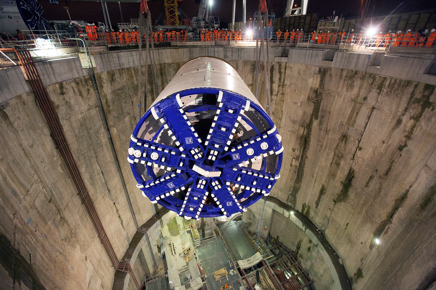 An enormous tunnel borrowing machine is suspended above a deep, round excavation, at night, surrounded by workers dressed in orange. 