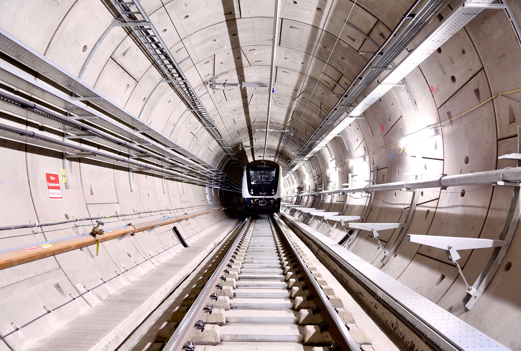 A sleek train approaches the camera in a white tunnel lined with power system equipment and other devices. 