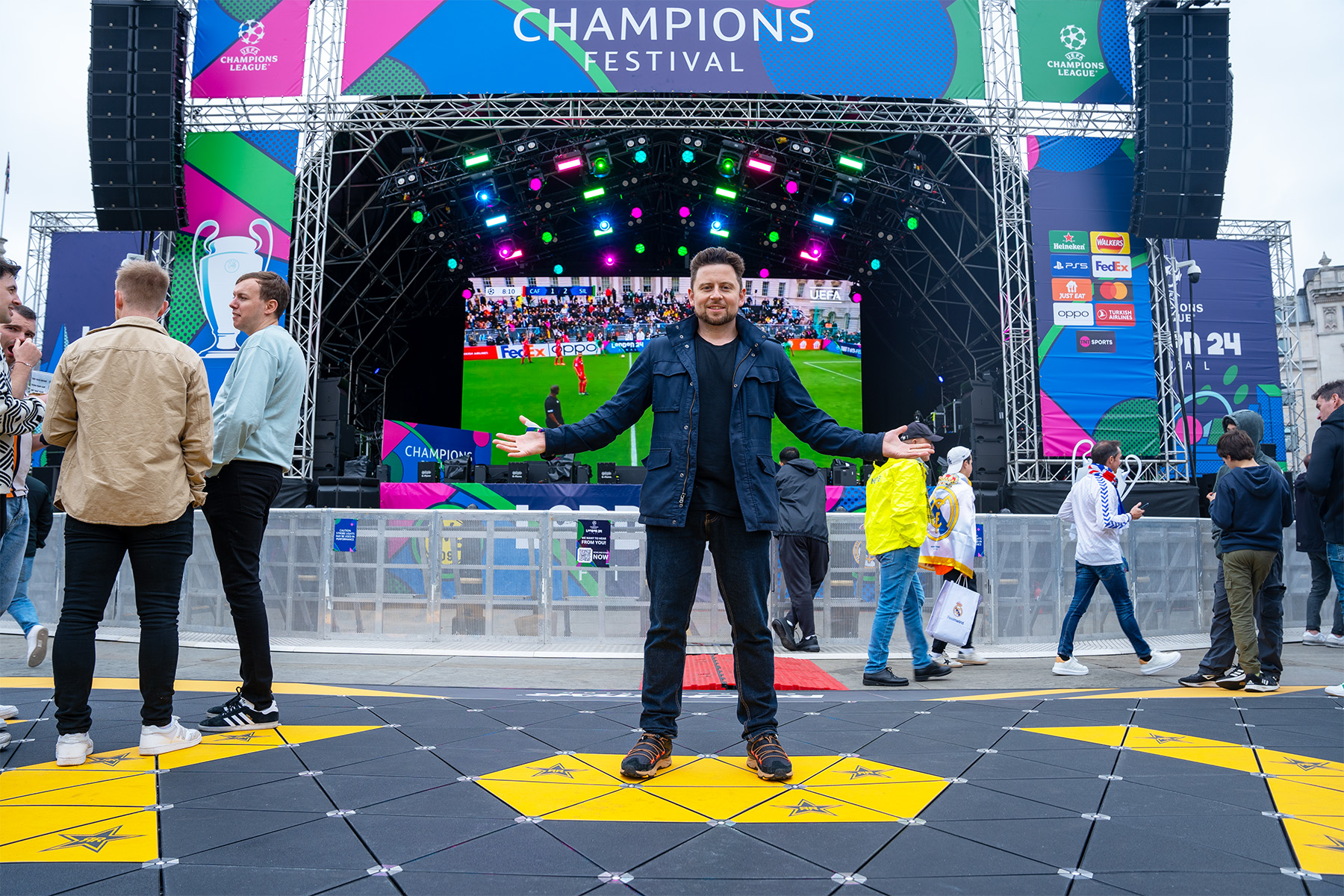 Image shows a man standing on a yellow hexagonal tile. There is a band behind him. 