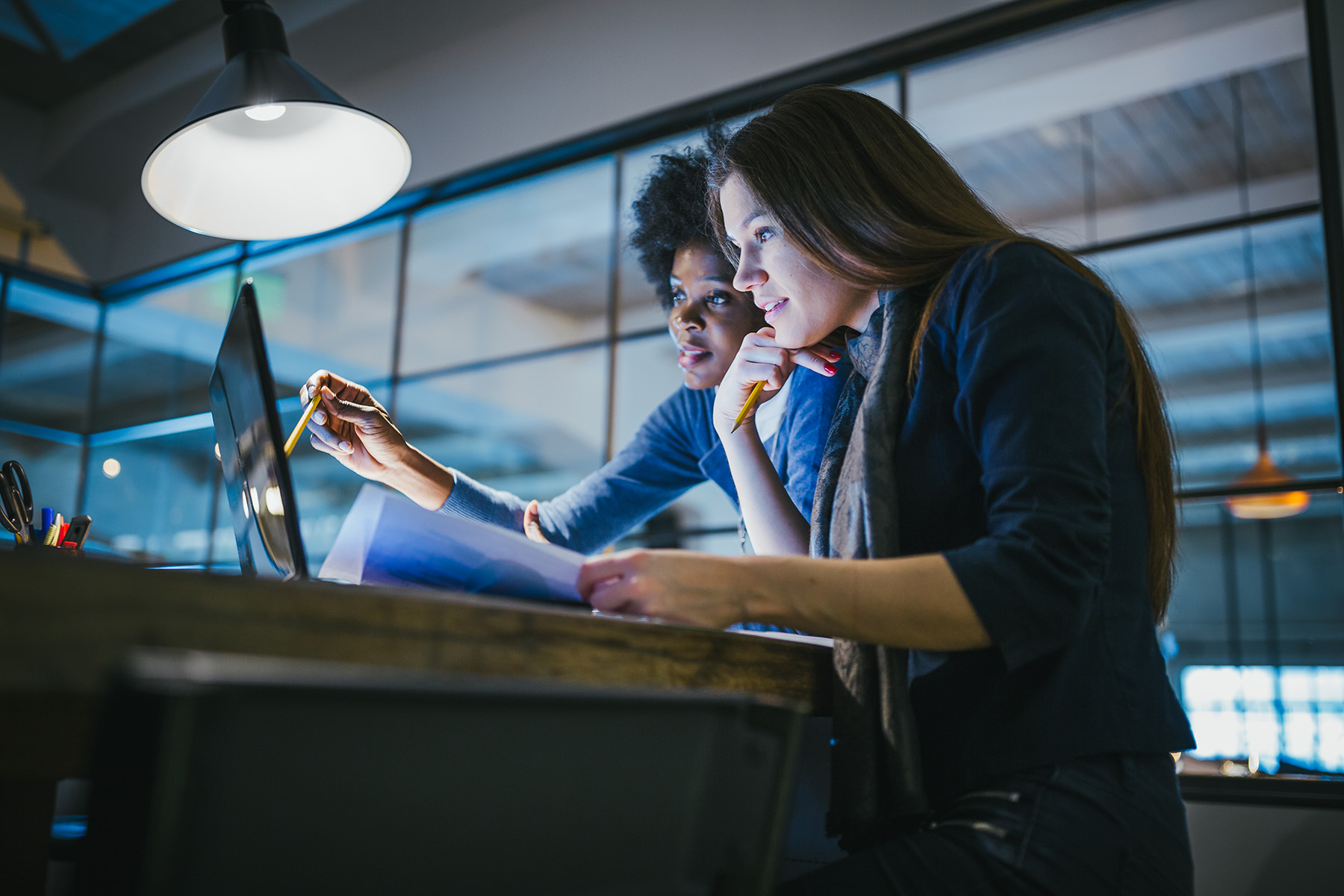 Two women at a workstation look at a laptop computer screen. 