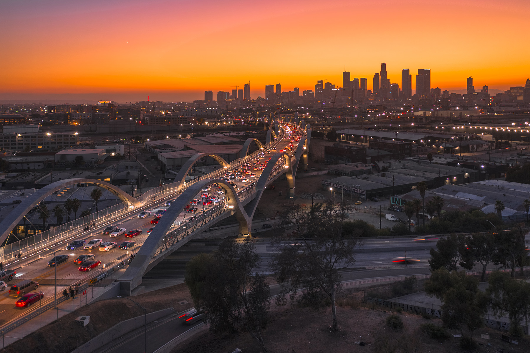 A four-lane bridge with arches stands above a Los Angeles highway. The city of LA is in the background, along with an orange sky. 