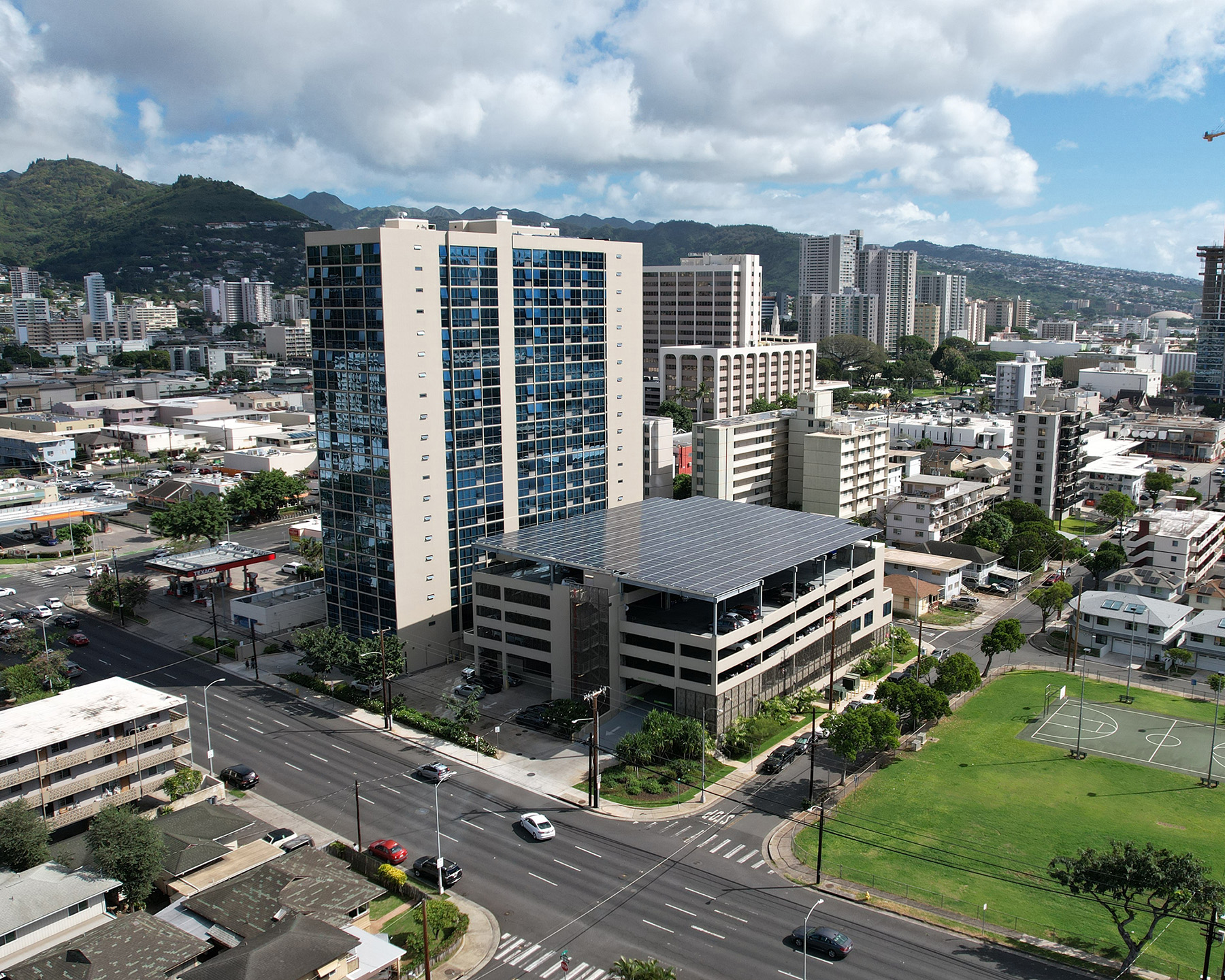 An apartment building towers over other shorter buildings and stands next to a 6-story parking garage with a solar paneled roof. 