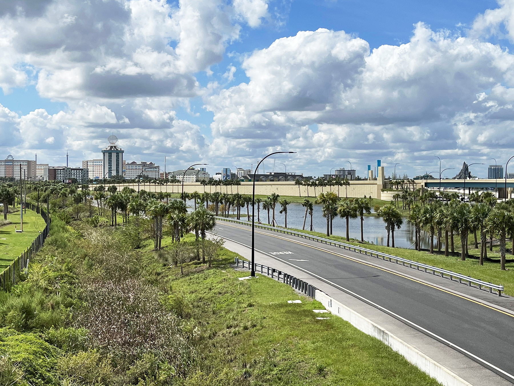 A new roadway is lined with palm trees. A blue sky with white, puffy clouds is in the background. 