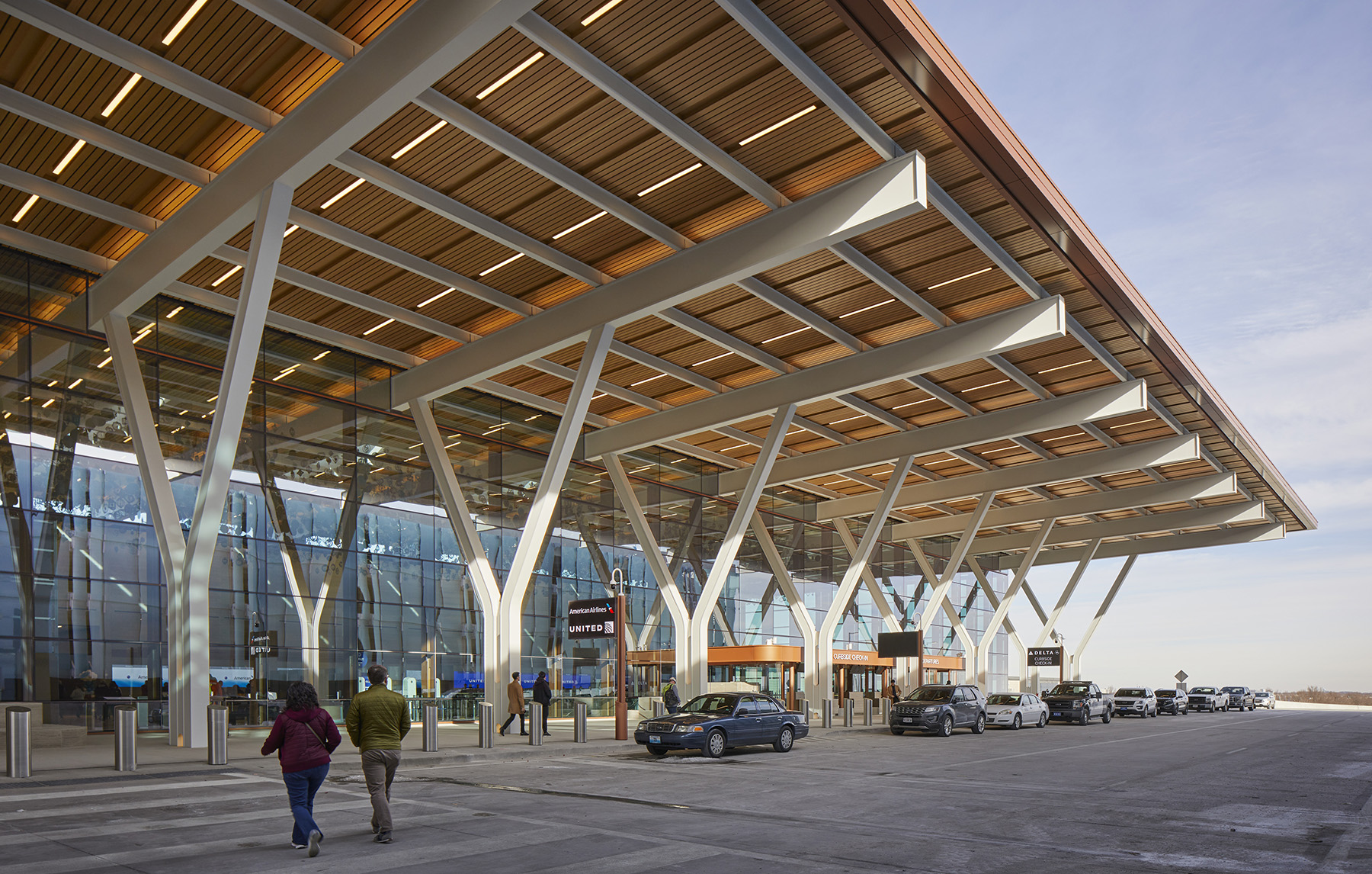 A large, tan-colored awning covers an airport drop-off zone. It is connected to a building in the background with a glass facade. 