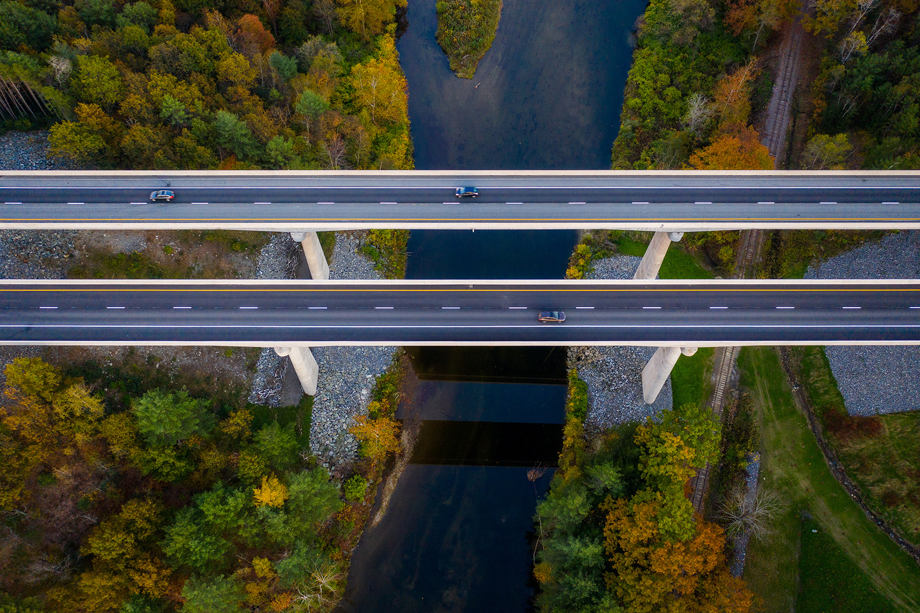 An aerial view of two parallel bridges, each with two lanes, tower over fall-colored trees and a small waterway. 