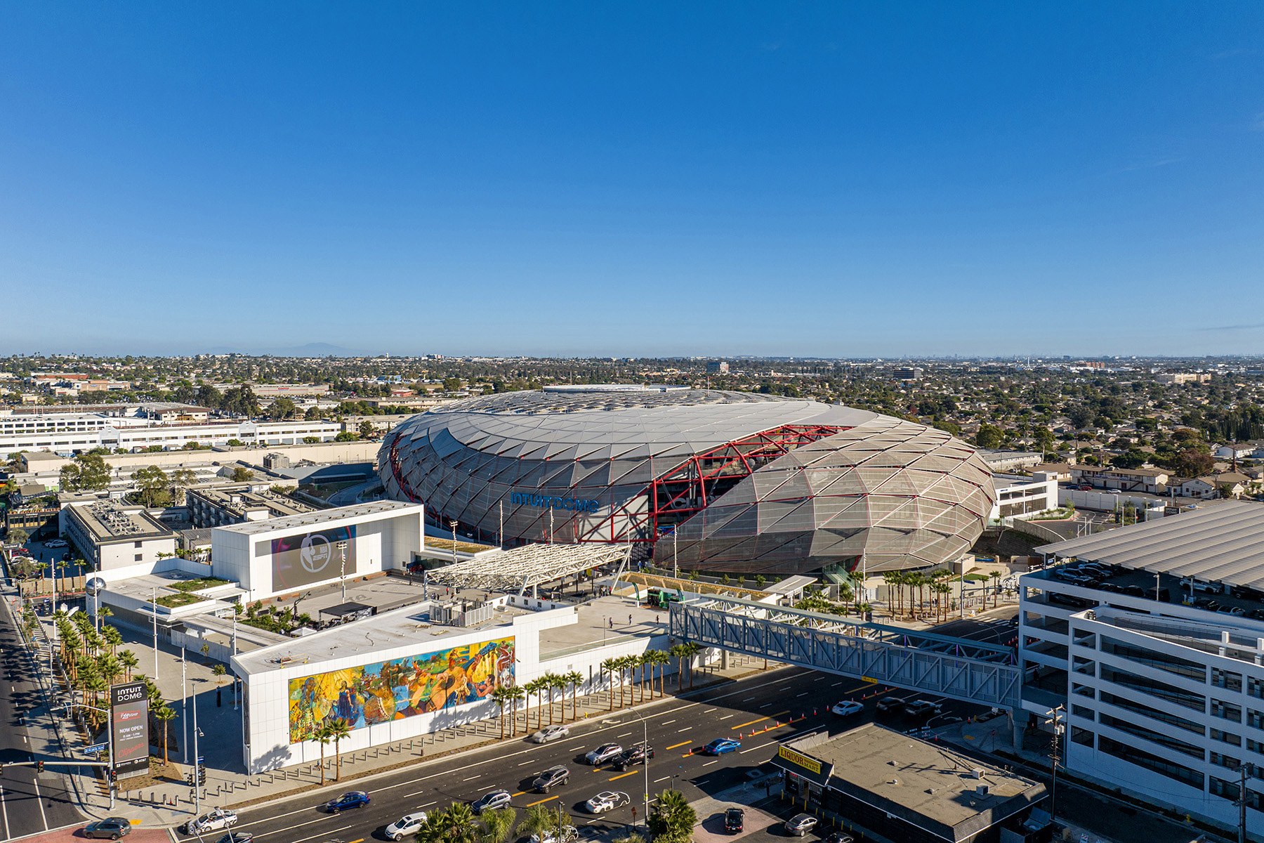 A sports arena sits amid the cityscape. There are walkways to the arena and many open entertainment and gathering spaces in front of the stadium. 