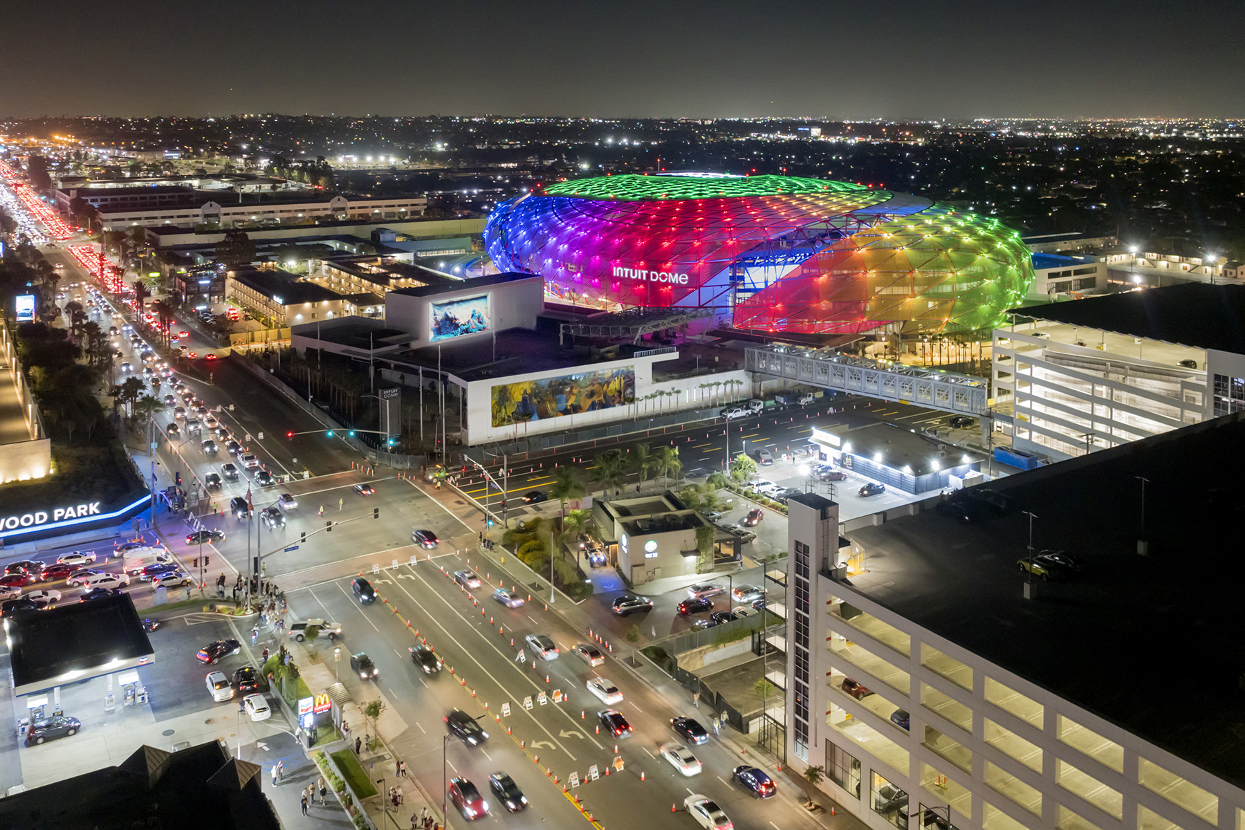 A stadium is lit up with lights of many colors. The walkway, parking garage, and other buildings can be seen in the foreground. 