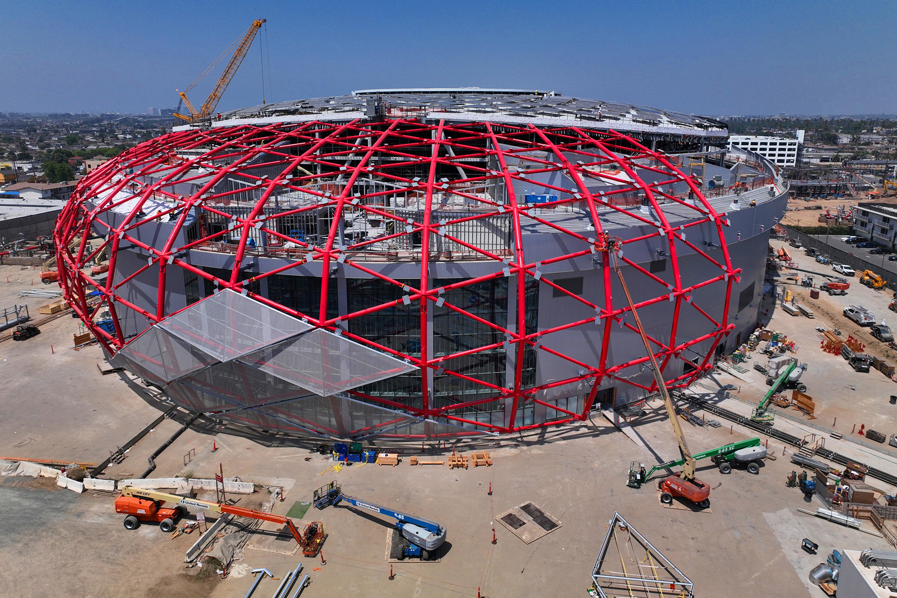 Image shows the diagrid shell of the Intuit Dome being assembled. Long red steel tubes are connected at various points. Mesh panels are being attached to the red tubes. 