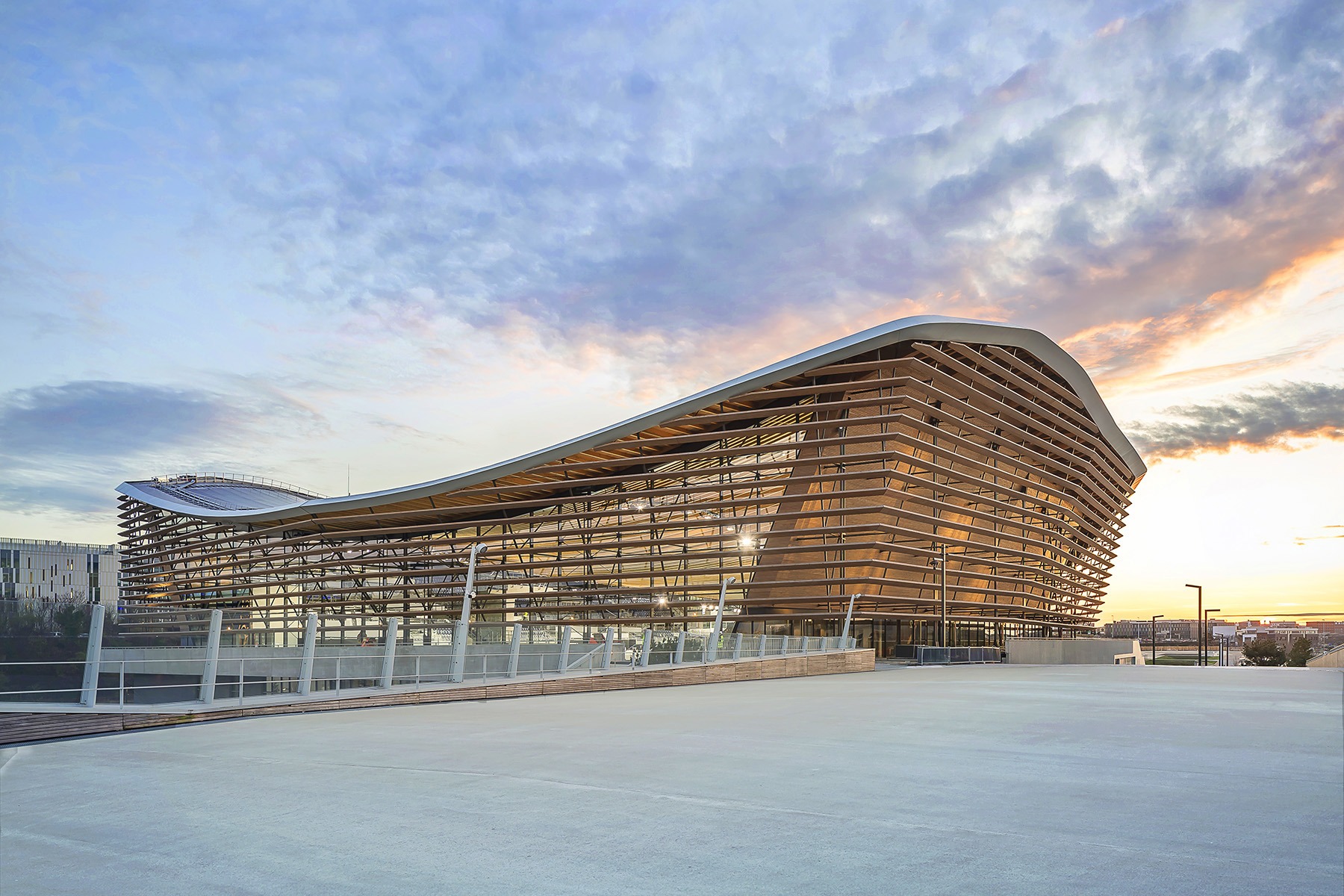 The finished building displays the timber elements comprising the sides and roof. A white pedestrian bridge is seen in the foreground and surrounding buildings in the background. 