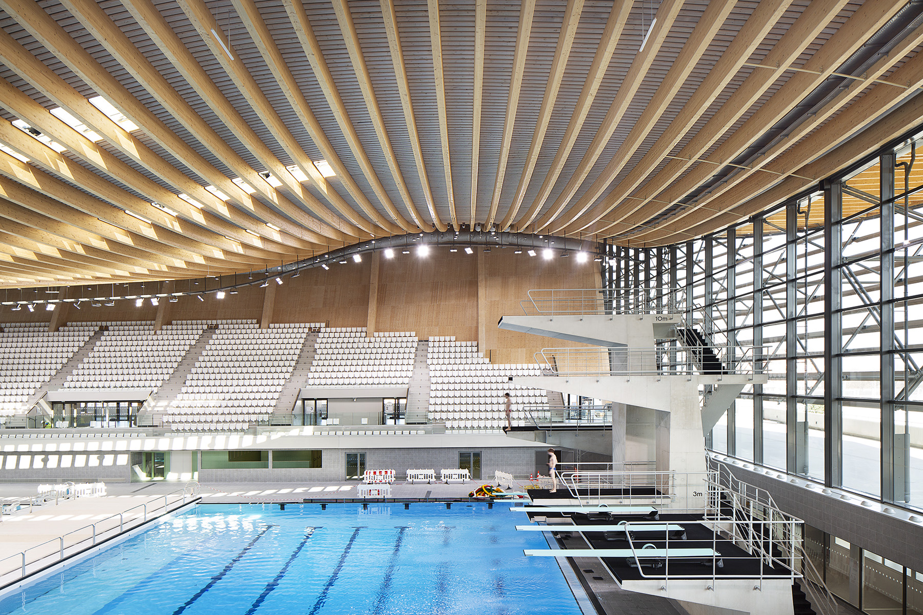 Five diving boards of various heights hover over the pool. White grandstand seats are to the right and in the background. 