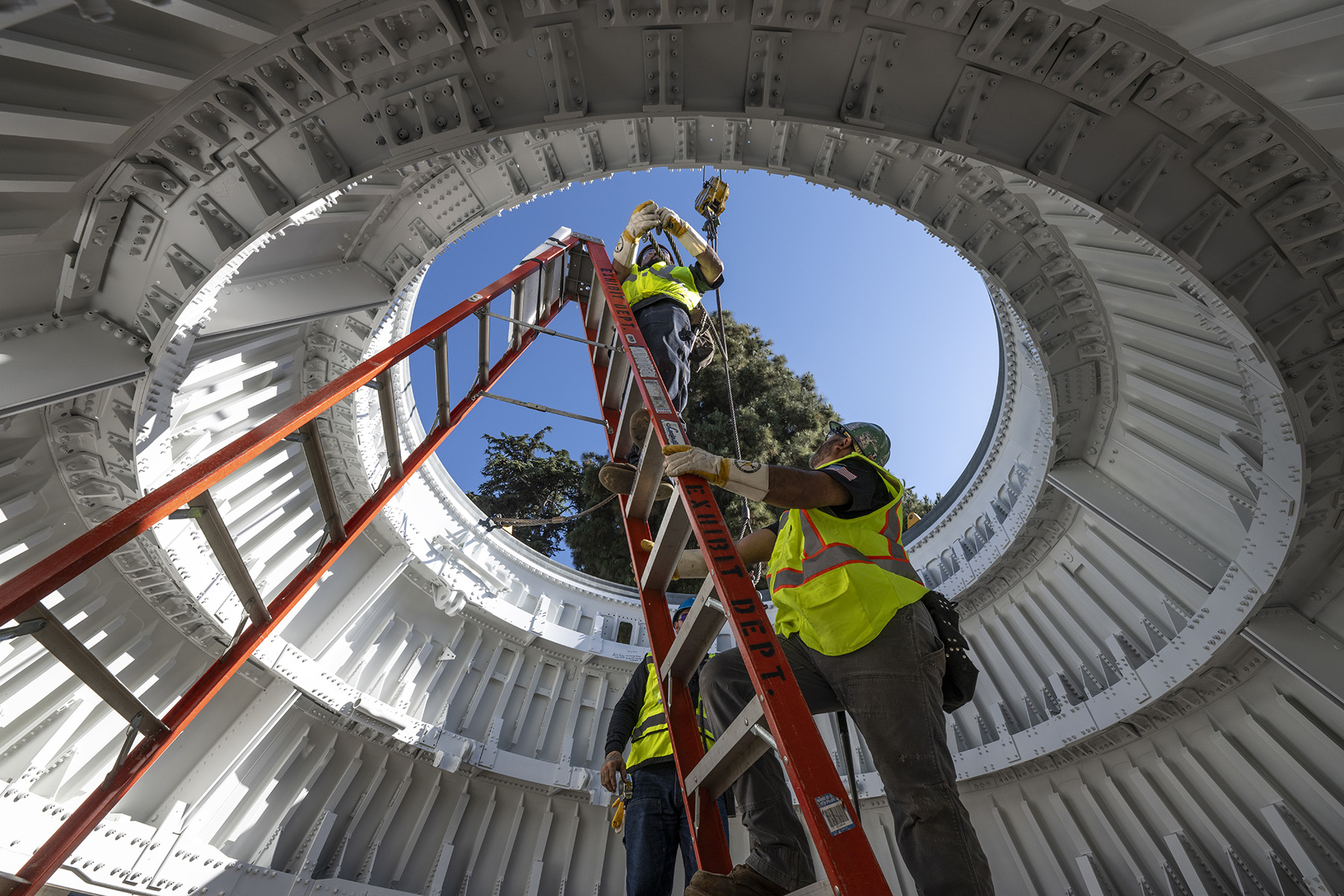 Workman in bright safety vests climb a ladder within the conical aft skirts. 