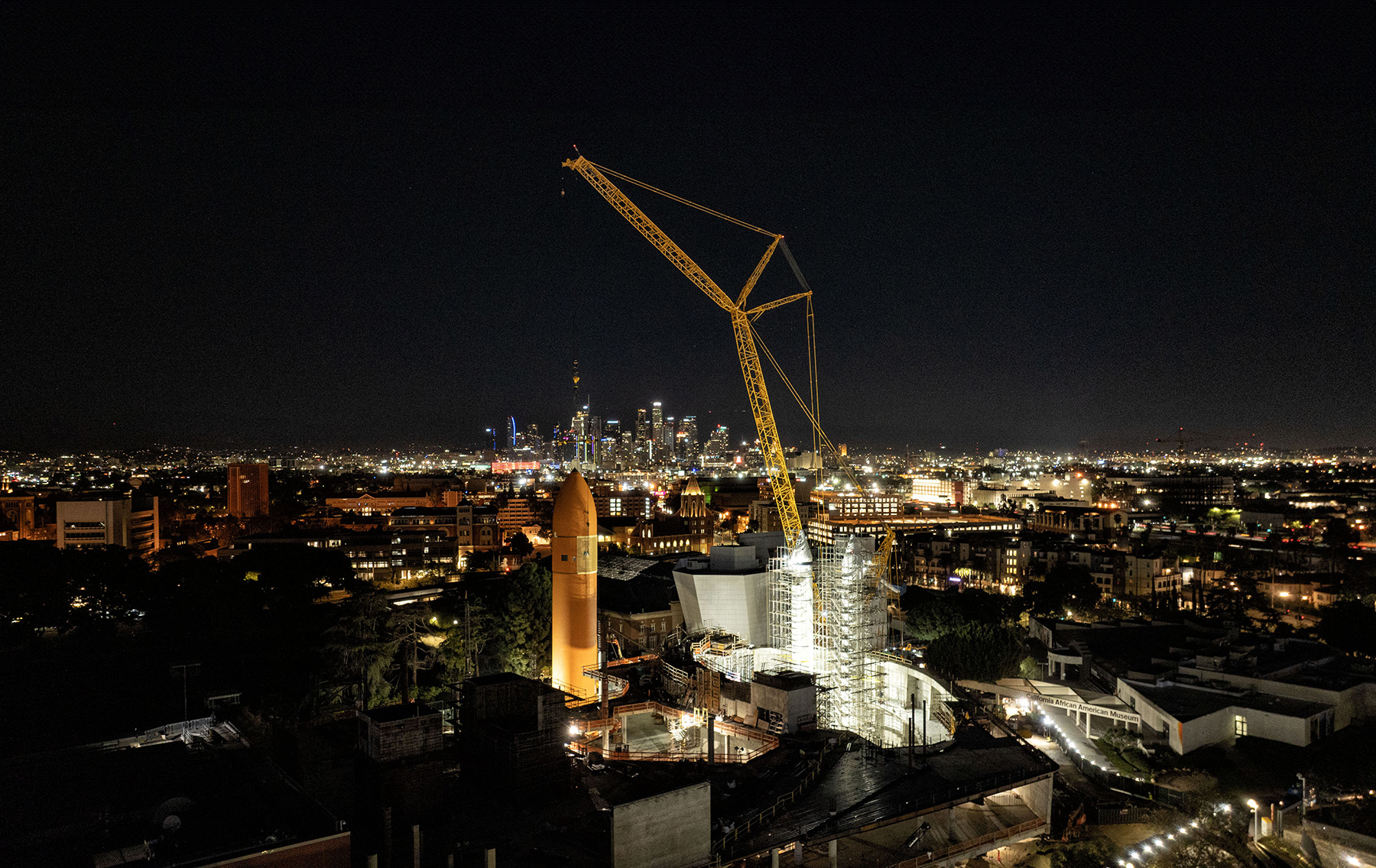 Towering cranes raise the orange external tank into position against a backdrop of the Los Angeles skyline at night