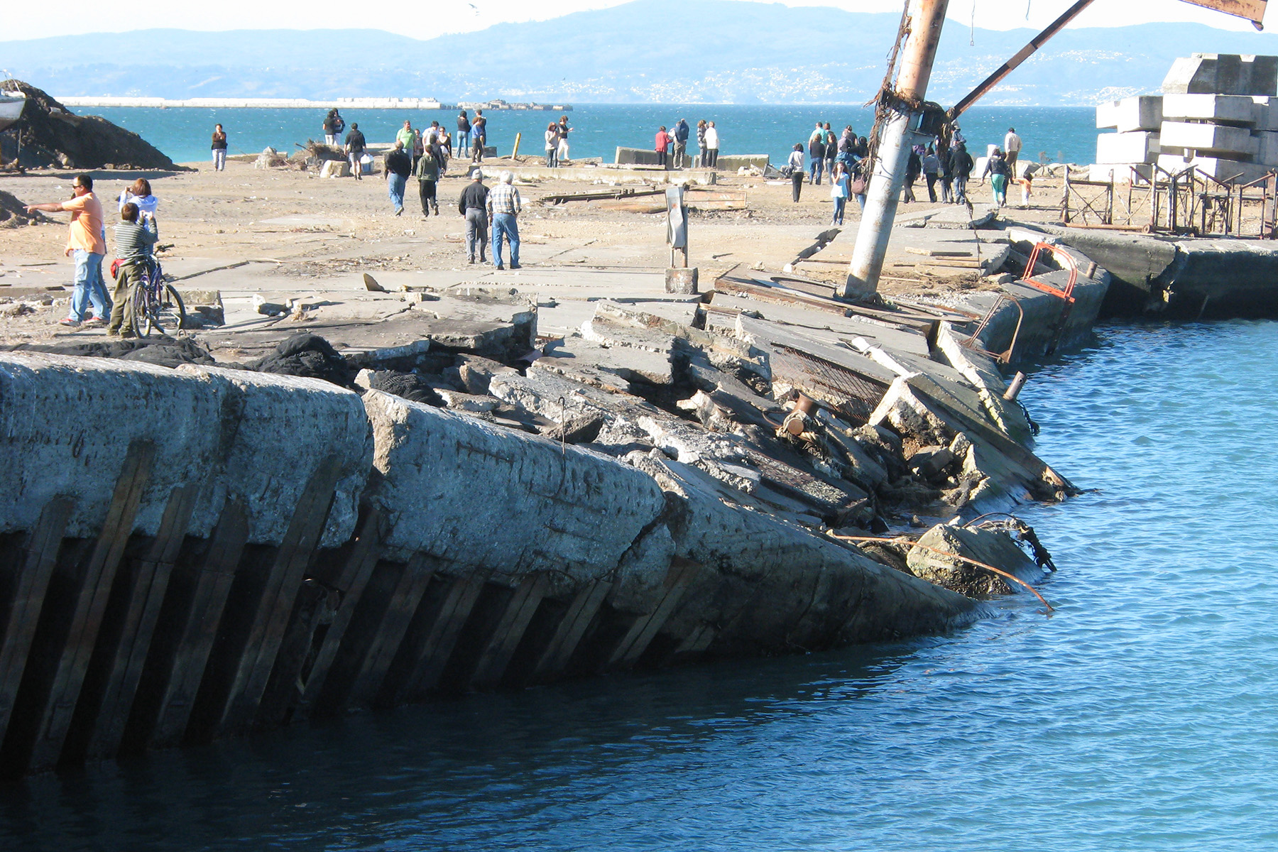 A retaining wall failed due to an earthquake and tsunami. People walk around on the pier.