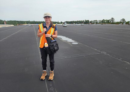 A smiling young woman, in safety vest and Atlas cap, stands atop the roof of a building.